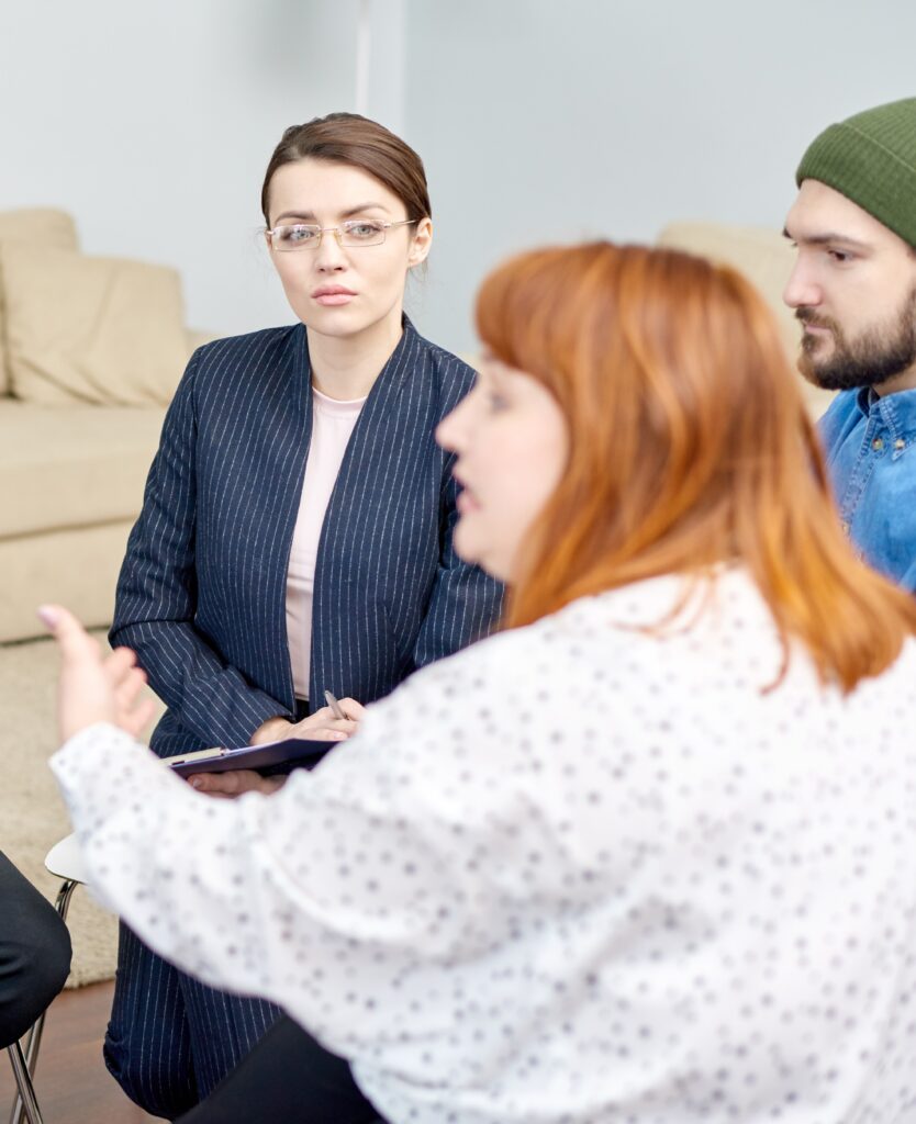 Profile view of red-haired obese woman discussing faced problem with other patients while participating in group therapy session, pretty psychologist listening to her with concentration