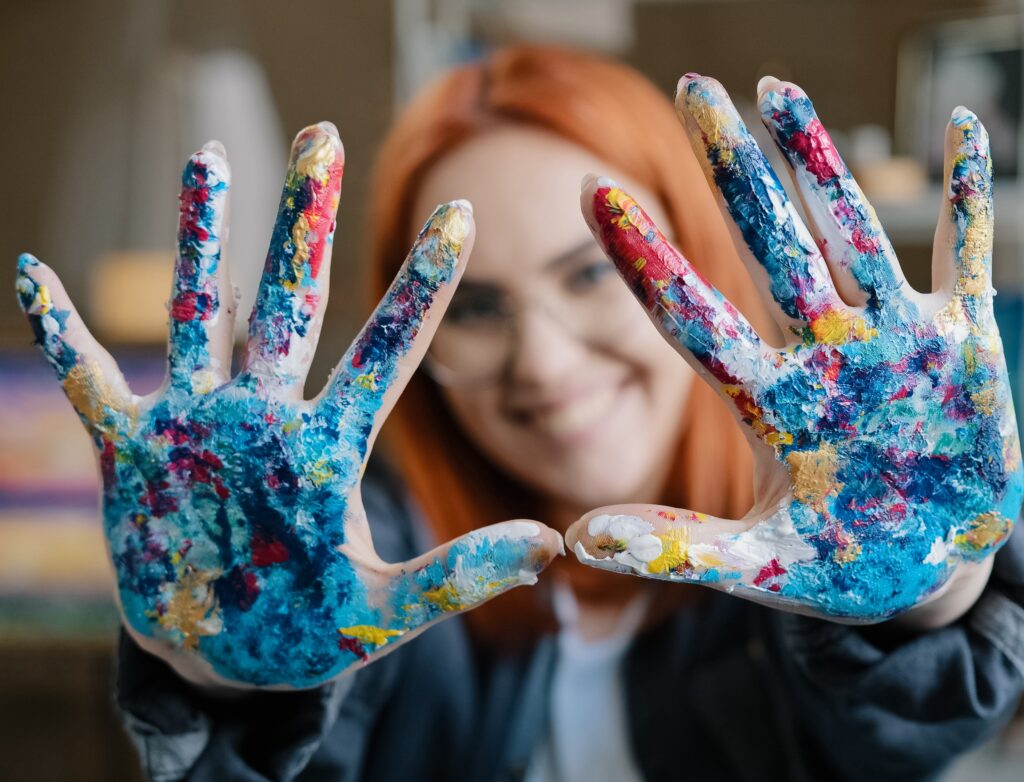Close-up happy funy woman with red hair painter girl artist in glasses looks at camera stretches forward hands showing dirty palms smeared in acrylic paint laughs inspired by drawing decor design art. High quality 4k footage
