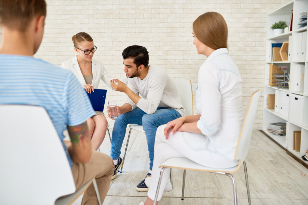 Portrait of young man crying in support group sharing problems with female psychiatrist and other participants in circle