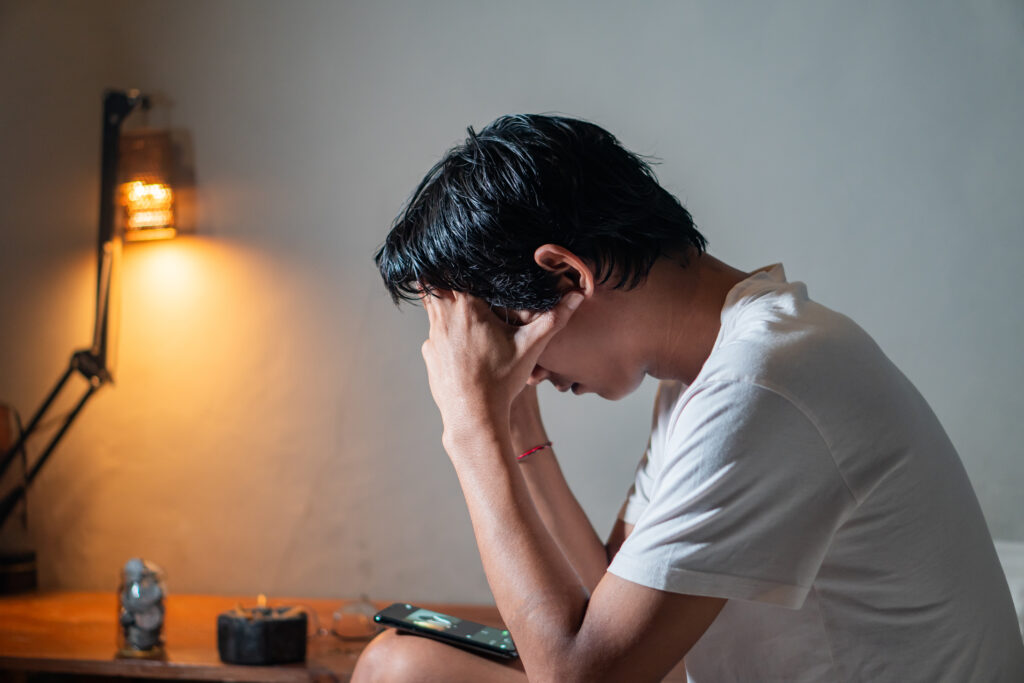 A person sitting on a bed with their head in their hands, appearing stressed or upset. A lit lamp and a smart phone are visible on the bedside table.